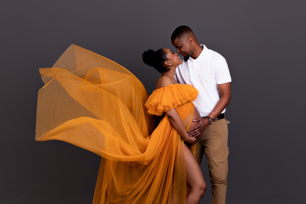Maternity photoshoot of an expecting couple with flowing orange fabric, capturing connection and partner involvement in Pretoria North birth support.