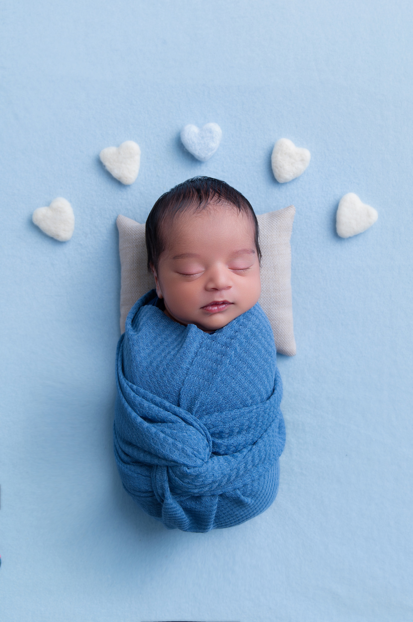 Wrapped newborn baby boy surrounded by white heart props on blue backdrop – newborn photoshoot Pretoria.