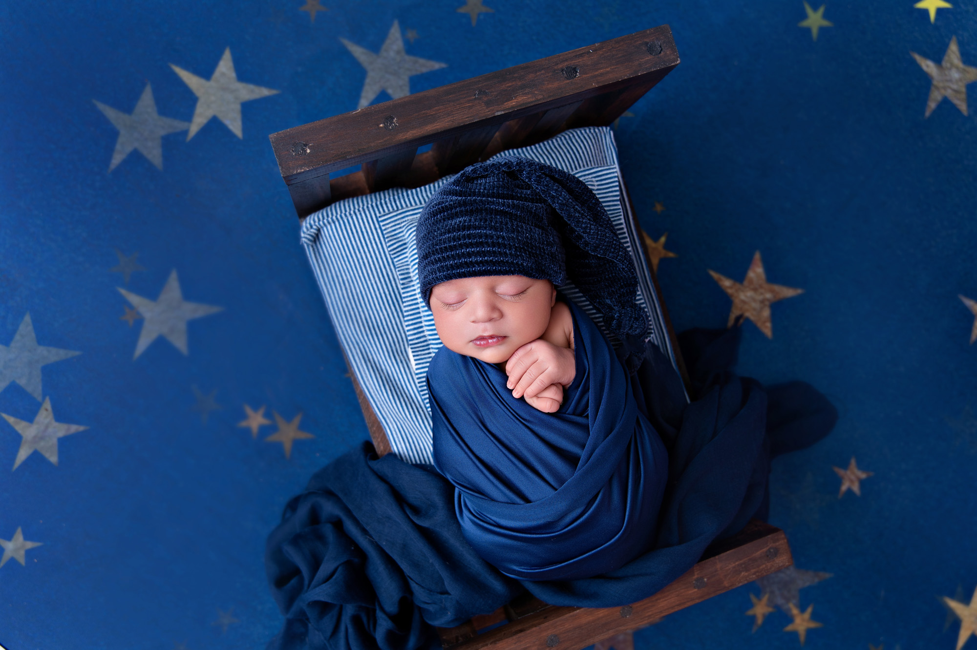 Newborn baby boy wrapped in navy blue on wooden prop with star background – newborn photoshoot Pretoria.