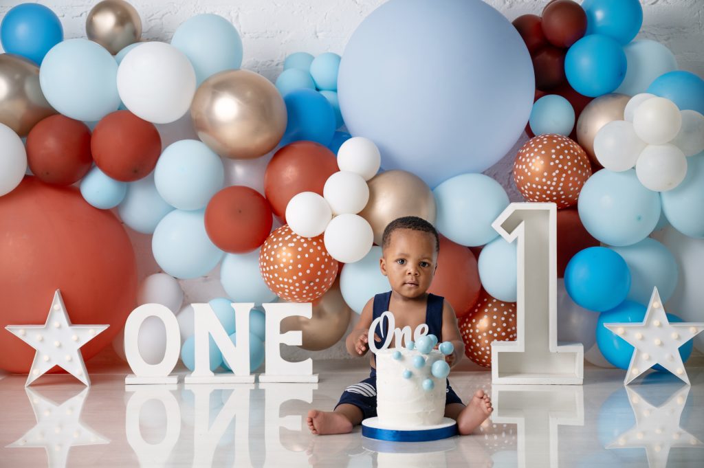 brown blue and gold balloons. baby boy dressed in pants with suspenders, smashing a cake