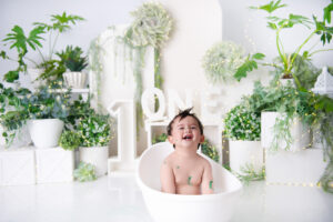 Happy baby sitting in white tub with greenery backdrop and “ONE” sign – 1st Birthday Cake Brits.