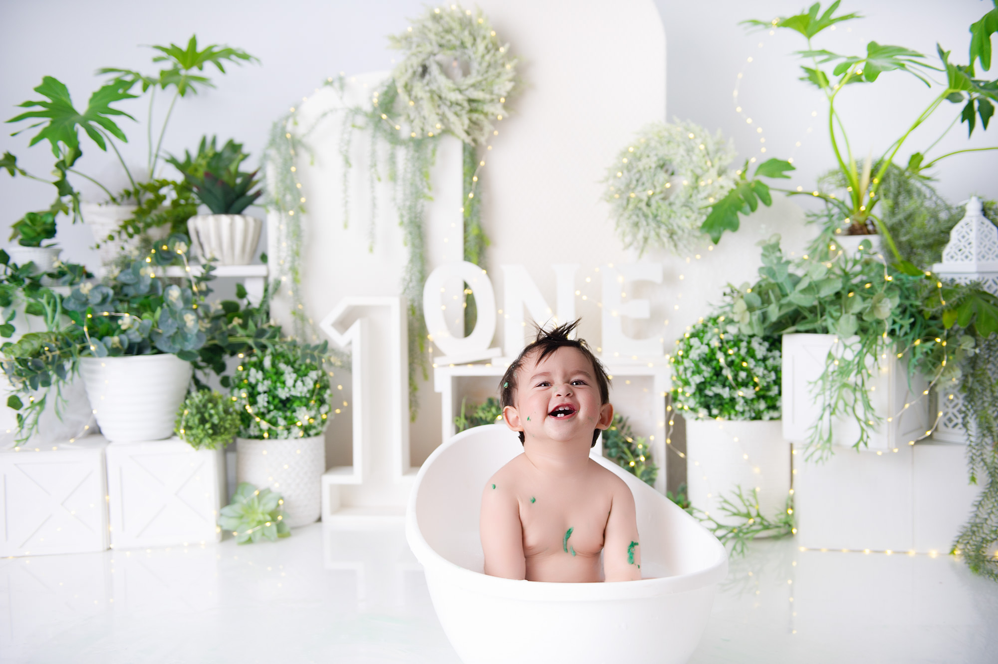 Happy baby sitting in white tub with greenery backdrop and “ONE” sign – 1st Birthday Cake Brits.
