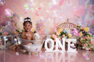 Baby girl laughing in bubble bath with pink balloons and “ONE” sign – 1st Birthday Cake Brits.