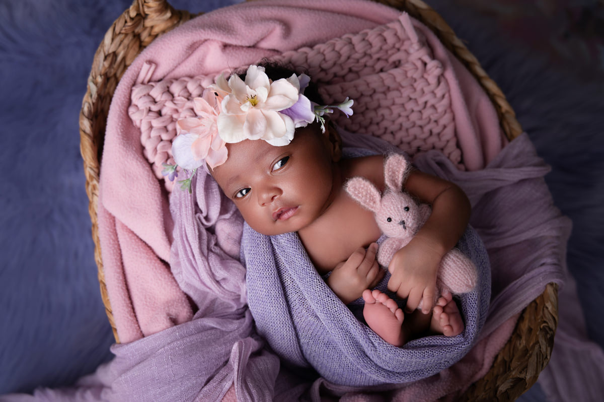 Newborn baby girl wrapped in soft purple and resting in basket with teddy during newborn week 1.