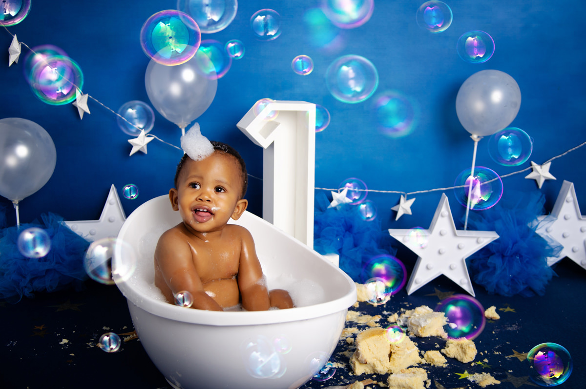 Bubble bath photo after cake smash with baby in white tub and number one backdrop – 1st birthday cake baker centurion.