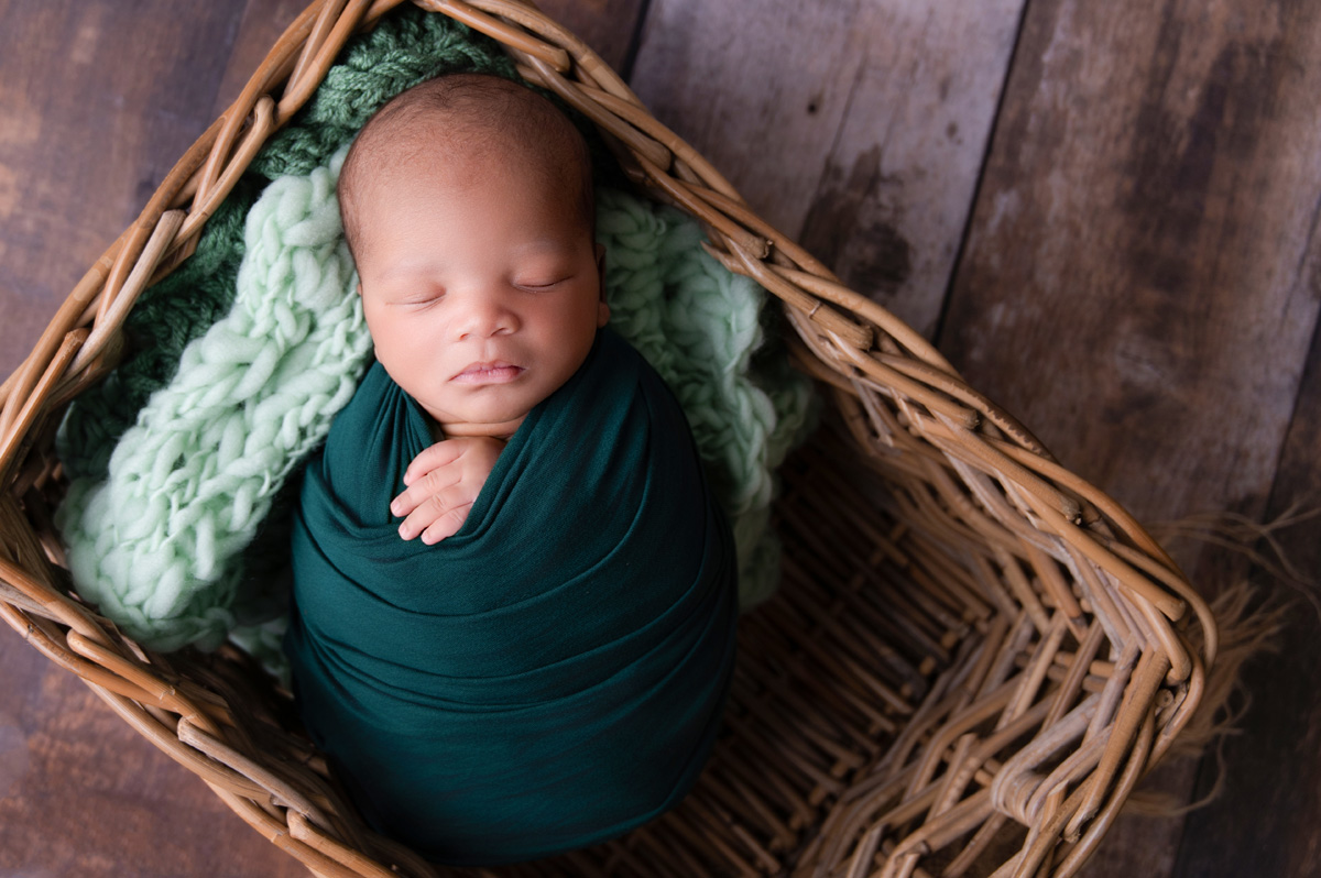 Sleeping newborn wrapped in deep green inside woven basket – Pretoria studio newborn photography.
