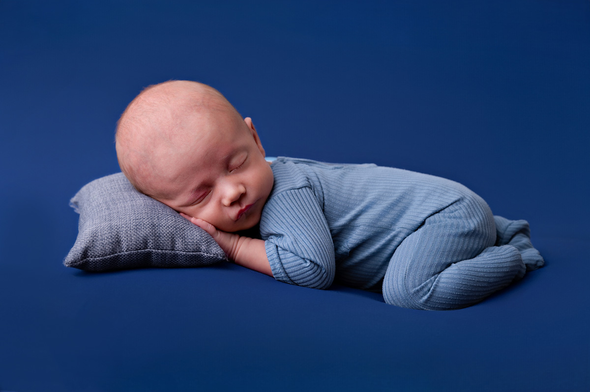 baby boy in a blue outfit sleeping on a soft blue backdrop.