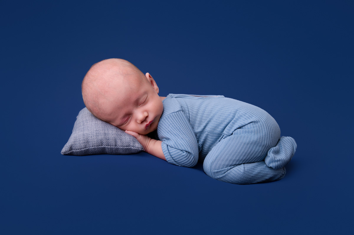 Newborn baby sleeping on a tiny pillow in a curled pose, styled in a serene blue setup