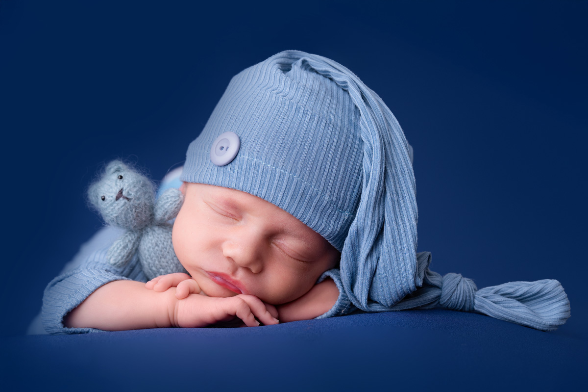 Newborn baby wearing a blue knitted hat, sleeping with a small toy, captured in a calm, safe newborn pose