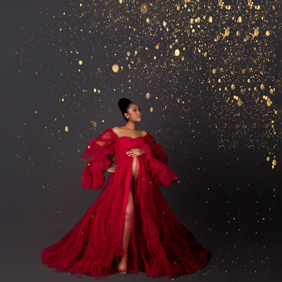 Pregnant woman wearing a dramatic red gown surrounded by soft golden sparkles, captured in a graceful studio pose